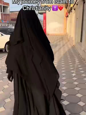 The image shows a person walking away from the camera, dressed in a long black hijab, in an urban setting. The person's back is turned, and they are walking on a paved pathway. The background features buildings and sunlight, suggesting an outdoor environment.