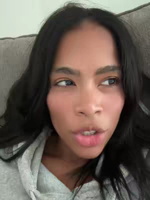 A close-up shot of a woman's face, looking off to the side. Her dark hair frames her face, and she has visible eye makeup. Her expression appears to be one of thought, slightly pursing her lips. The background is soft focus.