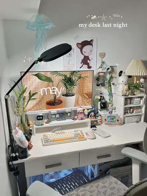The image showcases a well-organized and aesthetically pleasing desk setup, ideal for gaming or work. The desk itself is white, with a variety of items arranged on top. A large monitor displays what appears to be a nature-themed wallpaper. There are plants, including a potted plant and some bamboo shoots, adding a touch of nature. A mechanical keyboard with orange accents sits in the center, alongside a computer mouse. Decorative elements such as small figurines, a retro-style clock, artwork, and a jellyfish-shaped mobile, give the desk a unique and personalized feel. A desk lamp provides focused lighting while the overall composition creates an inviting and cozy ambience.