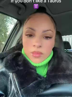 A woman is seated in a car, facing forward with a close-up shot. She's wearing a black fur-lined jacket over a bright green turtleneck. Her hair is pulled back, and she has makeup on. The background includes the car's interior, with indications of a window and possible outdoor scenery.