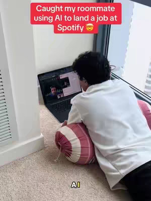 A person is sitting on the floor with their back to the camera, leaning against a large pillow, and looking at a laptop. The screen shows a video call with another person. Above the laptop screen, a red rectangle of text says, 'Caught my roommate using AI to land a job at Spotify'. The background includes a window and a light-colored wall.