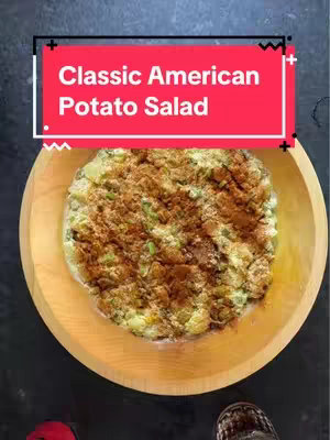 A top-down shot displays a wooden bowl filled with potato salad. The salad appears to be a mix of cubed potatoes, visible bits of celery and what looks like chopped green onions. It is also topped with a light dusting of spice, likely paprika or similar. The bowl is on a smooth, dark surface, and the image is well-lit with natural light. Overlaid on the top of the image is a rectangular text box with the words 'Classic American Potato Salad' in white sans-serif font.
