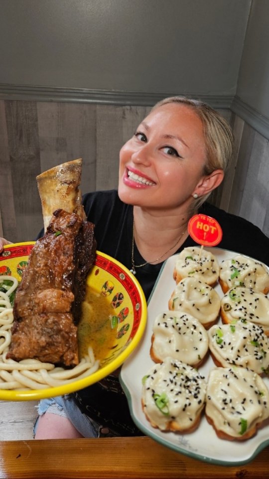 A woman with light blonde hair and a black shirt smiles, holding a plate of food in each hand. On the left, a plate of noodles with a large beef rib, and on the right a plate of dumplings. A red 'HOT' sticker is by the dumplings.