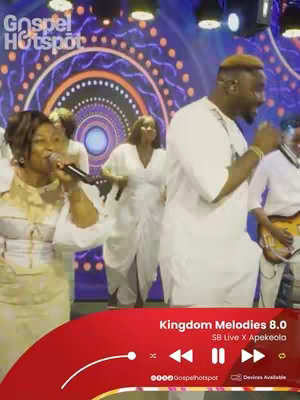 The image displays a live performance of a gospel music concert. Several people are on stage: a male singer with a gold-colored hairstyle is central to the image, singing into a microphone and is surrounded by female and male singers. The background has a light show. A red bar at the bottom has the music title and playback controls, indicating a media player interface. The Gospel Hotspot branding is present.