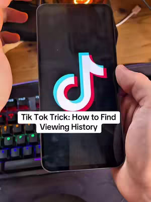 A hand holding a black smartphone displays the TikTok logo on a dark background. Overlayed at the bottom is a white box with black text that reads 'Tik Tok Trick: How to Find Viewing History'. Background elements include a keyboard, desk and other objects appearing out of focus.