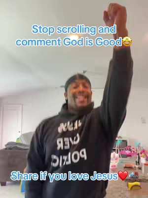 A man with a black durag and black hoodie overlayed with text, raising his right arm up in a celebratory gesture, smiling at the camera. The background contains a living room with objects and pink toys and other decorations. The video is shot in portrait mode. 