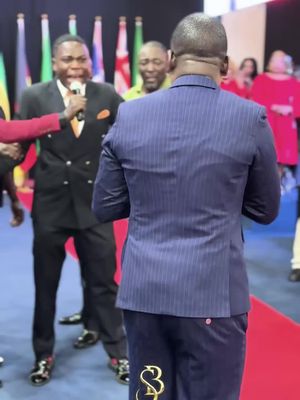 The image captures a scene at what appears to be a religious event or presentation. The main focus is on a man’s back wearing a navy blue pinstripe suit. In front of him, a man in a black suit with an orange tie and a microphone, is addressing an audience. Other people are present, and flags are visible behind them, indicating an international event. A red carpet adds a formal tone to the setting.
