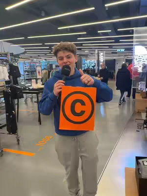A young man, holding a microphone in his right hand and an orange sign with a black "C" inside a black circle in his left hand, gives a thumbs-up with his right hand, in a modern retail store.