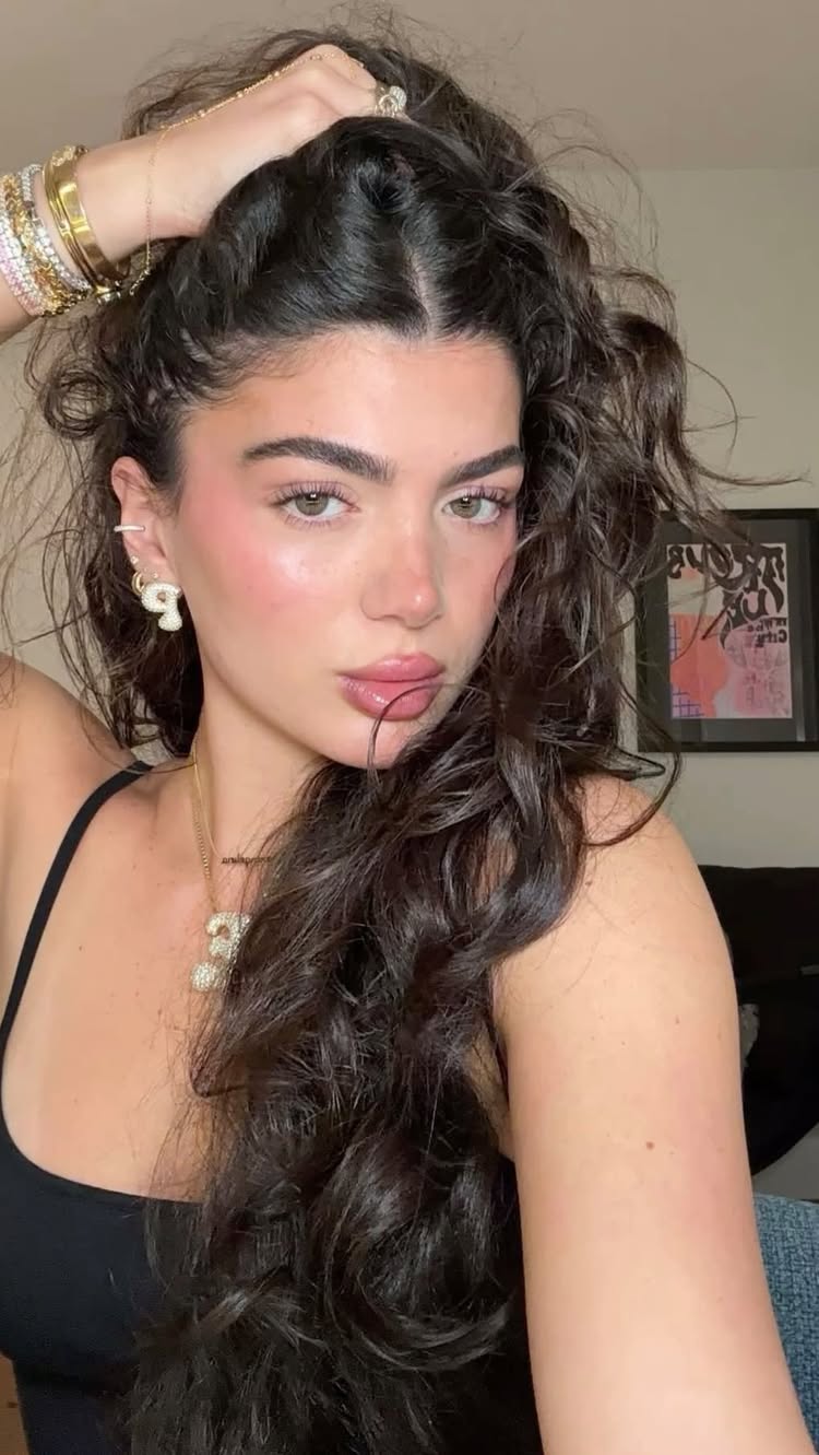 A selfie of a woman with long, dark, wavy hair, wearing a black tank top. She has full lips, flushed cheeks, and rosy pink coloring around her eyes. She has multiple gold bracelets and rings and a gold necklace. The background shows parts of her room with a picture frame.