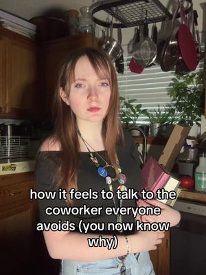 A woman with long brown hair and fair skin looks at the camera with a neutral expression. She is wearing a black off-the-shoulder top and a necklace of colorful beads. She is standing in a kitchen, with a countertop behind her that has various items on it. Text overlayed on the bottom of the image reads 'how it feels to talk to the coworker everyone avoids (you now know why)'.