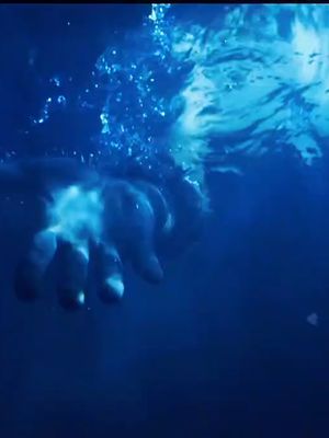 An underwater scene with a hand reaching upwards towards the surface. The water is a deep blue, and the hand appears partially illuminated, possibly by light filtering from above, with bubbles and water droplets.