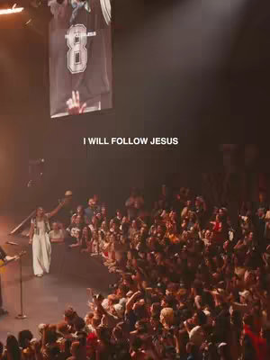A live performance featuring a female singer on stage in front of a large crowd. Text overlay reads 'I WILL FOLLOW JESUS'. The background is blurred, with stage lighting adding a warm glow.