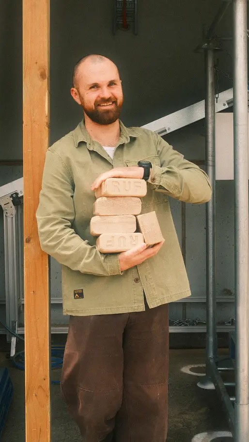 A man with a beard, wearing a green shirt and brown pants, stands smiling and holding a stack of light-colored rectangular briquettes. The background has a simple construction setting, with a partial shelter and metal supports visible.