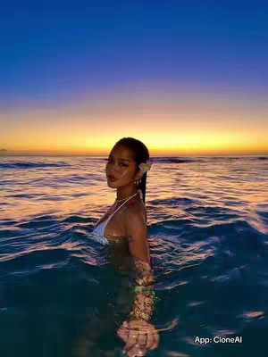 A woman is standing in ocean water, seen from the chest up. She has dark hair, a flower behind her ear, and is wearing a white bikini top and a necklace. The water is a blend of blues at the bottom and transitioning to orange and yellow at the sunset in the top of the image. The AI app logo is at the bottom right.