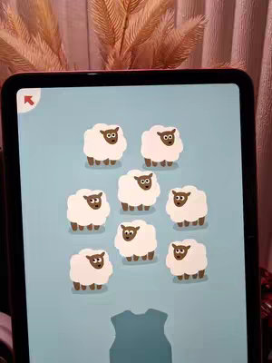 An iPad screen displays a game scene with a light blue background. Nine fluffy white sheep with brown faces and legs are scattered across the screen. In the lower part of the screen, there is a dark bluish silhouette of what appears to be a piece of clothing. The background is blurred and has pink and brown tones, possibly from surrounding decor.