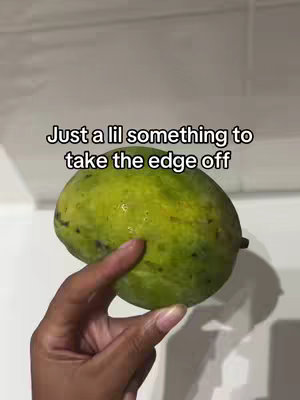 A close-up shot features a hand holding a large, oblong green mango against a plain white background. The mango appears fresh and slightly wet. Above it, text overlays read, 'Just a lil' something to take the edge off.'
