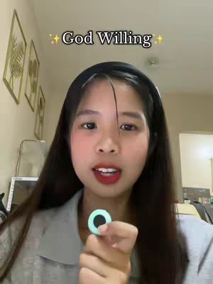A young woman with long dark hair smiles directly into the camera, holding a circular teal object. The background shows a typical home interior with wall decor. Text overlayed on top reads 'God Willing' with sparkles.