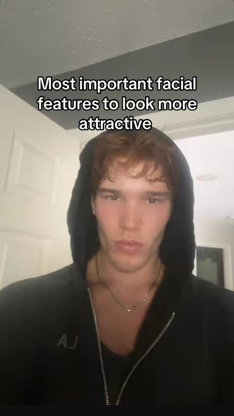 A young man with light skin is centered in the frame, wearing a black hoodie with the hood up, against a background that appears to be a lightly textured wall. Text overlays the top stating, "Most important facial features to look more attractive." The man is looking at the camera with a neutral expression. The composition is simple, focusing primarily on the man's face and upper body.