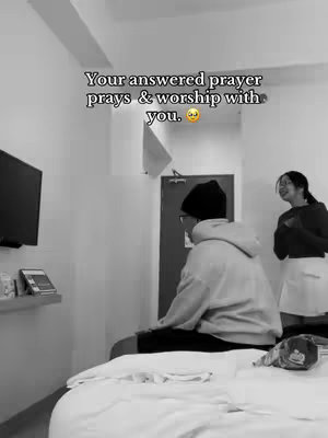 A black and white photo shows a man sitting on a bed, turned away from the camera. A woman stands in the background, smiling and gesturing. Text is overlaid at the top. It appears to be a hotel room setting.