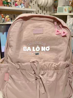 The image features a close-up of a light pink backpack. The backpack has a textured, slightly quilted appearance. Centered on the backpack are the words "unboxing" and "ba lô nơ" in a playful font with some stars. It is set against a background that suggests a room, with items visible like shelves and decor.