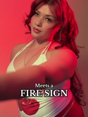 A woman with red hair poses against a red backdrop. She wears a white top with a cherry-like design and a necklace. Text overlays read 'Meets a FIRE SIGN'.