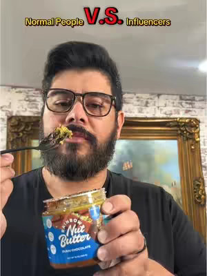 A man with glasses and a beard is shown taking a bite of food from a fork, holding a jar of Rainbow Nut Butter. The background includes an ornate painting and a partial brick wall.