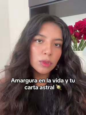 The cover image showcases a medium shot of a woman with dark hair looking directly at the camera. She has fair skin and is wearing a dark-colored top. Behind her, a vase contains red roses. Overlaying her image is white text that reads "Amargura en la vida y tu carta astral," with a small graphic of a planet or star next to the text.