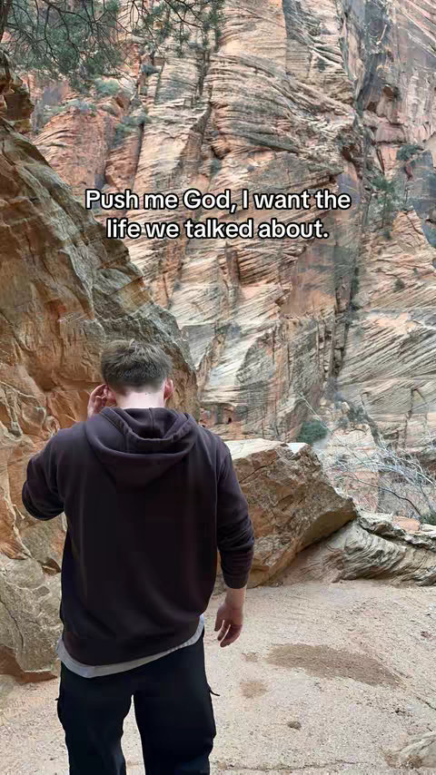 A man stands with his back to the camera, facing a rugged canyon landscape. Text overlays the image. The man is wearing a dark hoodie and black pants. The background features tall, striated rock formations in shades of brown and orange, with hints of green vegetation visible. The sky is out of view.