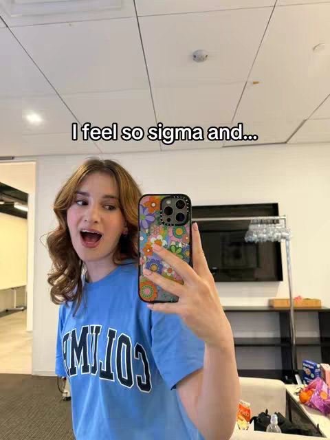 A young woman is taking a selfie in what appears to be an office or studio setting. She is wearing a blue t-shirt with white block letters, and her long hair is styled in loose waves. She holds her smartphone, which has a colorful floral case. The background is blurred, with what seem to be office furnishings and a white ceiling visible.