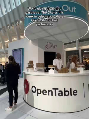 The image captures a pop-up event at The Oculus, a modern architectural space. The foreground features a counter branded with 'OpenTable' and a red graphic. Behind the counter, two individuals, one male and one female, are serving or interacting with customers. A banner overhead indicates 'Give Out' and 'Little Ruby's'. The scene is well-lit, suggesting a vibrant event. In the background, other attendees are visible, adding to the atmosphere.