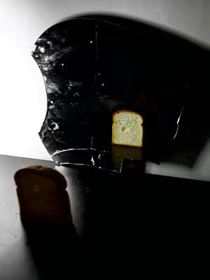 A studio shot featuring a slice of bread placed on a dark surface, with a contrasting backdrop. The bread slice is illuminated, casting shadows and highlighting its texture. The composition is minimalist, focusing on the subject's form and contrast.