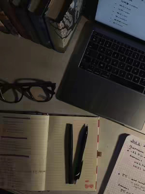 A top-down shot of a study desk with a laptop, a stack of books, glasses, and study materials. The warm lighting sets a mood, with the open notebook and pens suggesting preparation and productivity.