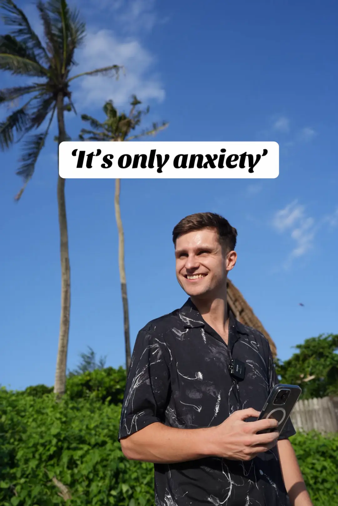 A man with short brown hair smiles while standing against a bright blue sky, palm trees, and green foliage create a tropical background. The man wears a black patterned shirt, holds a phone, and a text overlay reading 'It's only anxiety' is in the top center of the image.