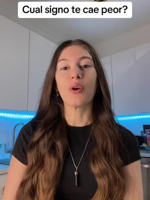 A young woman with long brown hair stands in front of a kitchen, her mouth open in a speaking position. Text in white overlayed on the top says, "Cual signo te cae peor?" with the background appearing to be a simple kitchen cabinet. There appears to be a necklace around her neck.