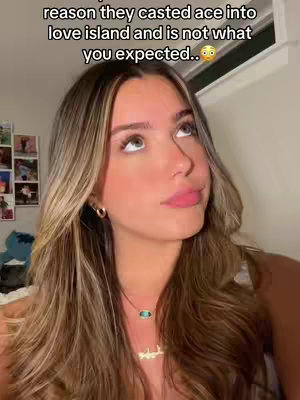 A young woman with long, wavy blonde hair looks upward with a contemplative expression. She wears a necklace and small hoop earrings. The background is a simple white wall with a partially visible window and a few polaroid photos.
