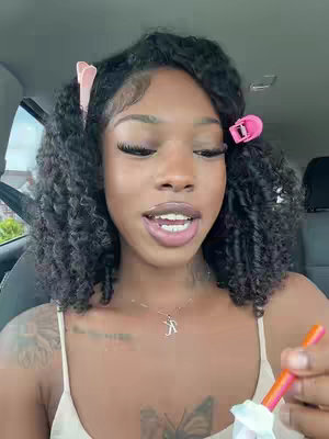 A young woman with curly black hair, styled with pink clips. She is in a car, looking toward the camera with a slight smile and holding a colorful orange drink. There are visible tattoos on her arm and a small necklace visible.