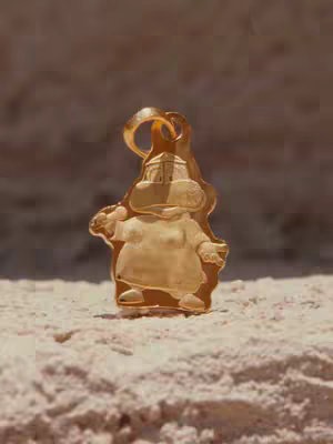 The image is a close-up of a gold pendant or charm. The charm is in the shape of a stylized cartoon character, with a large body, small arms, and a head featuring design elements. The character is standing on a textured, light-colored surface that appears like fine sand or a similar material. The background is blurred and of a brown color, possibly a wall or a textured backdrop, this enhances the gold pendant's prominence.