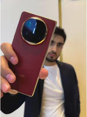 A man holds up a maroon smartphone with a large circular camera module featuring a lens and additional lighting. The background is blurred, with hints of a white wall and a dark blazer. The man is looking at the camera with a serious expression.
