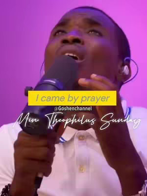 A medium shot of a Black man singing into a microphone. He has a white polo shirt on and black beaded bracelets. A yellow bar overlay has the text "I came by prayer" with another line of white text below it. A white cursive font is used along the bottom of the image with the name "Min Theophilus Sunday". The background is a blurred combination of pink and purple.