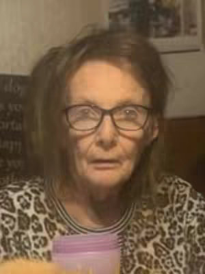 The image shows an older woman with glasses looking directly at the camera. She has short, light-brown hair and wears glasses. Her shirt has a leopard print-like pattern. In the foreground, a portion of a container can be seen.