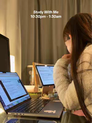 A woman is shown from mid-back, engrossed in focused study. Two laptops are open on a desk, revealing screens with what appears to be documents or notes from the perspective of the viewer. At the top of the image, centered, the overlaid text reads 'Study With Me, 10:30 pm - 1:30 am'. The lighting is dim and warm, casting a soft glow over the scene, creating a cozy atmosphere.