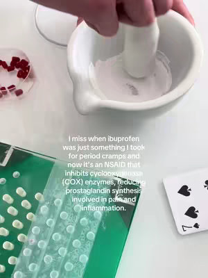 The image shows a close-up of a person's hand using a pestle to grind something in a mortar. To the right is a deck of cards with the 7 of spades visible. To the left is a tray with pills of the same color as the hand's nail polish. The lighting is bright and sterile, typical of a lab setting.