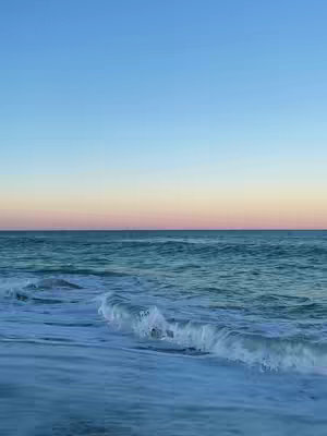 An ocean scene at sunset. The sky transitions from a deep blue at the top to a soft pink and yellow near the horizon. The ocean waves gently roll toward the shore, creating a peaceful and serene atmosphere.