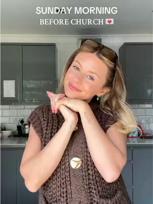 A woman with blonde hair and sunglasses on her head is smiling, with her hands folded under her chin. The background is a kitchen with cabinets and appliances.