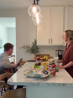 A man is playing the guitar while two people are in a kitchen setting. White cabinets are visible with a countertop that has some food items. The room has bright lighting, with a light fixture visible.