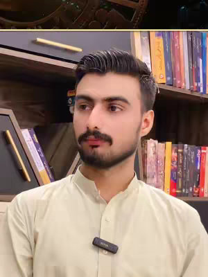 A man with dark hair and a mustache is centered in the frame, facing slightly to his right. He wears a light-colored collared shirt and a small microphone is clipped to it. A bookshelf filled with books forms the background. The composition has good lighting, highlighting the man's face.