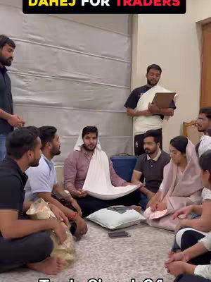 A group of people are gathered in what appears to be a living room, around a central figure draped in a white cloth. They are looking down at the figure, who is the main subject of the image. Above, in a red banner, the text
"DAHEJ FOR TRADERS" is displayed, indicating a humorously-themed skit. The lighting seems natural, with many people seated, and the setup indicates an event.