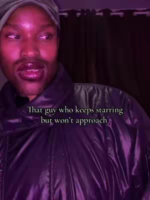 A man with a subtle mustache and stubble is looking to the side, likely in a studio setting. The background shows a dark curtain. The man wears a black quilted jacket and a dark gray beanie style hat. The primary color is pink due to the artificial lighting. Text overlay saying 'That guy who keeps starring but won't approach' is added to the cover image.
