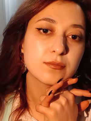 A selfie-style shot of a woman with auburn hair and subtle makeup, looking directly at the camera with a contemplative expression. She is holding her hand up to her chin. The lighting seems to be soft, possibly from a window, giving a warm glow to the scene.
