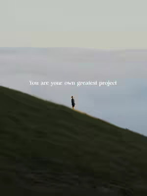 The image depicts a person standing on a green hillside, silhouetted against a backdrop of layered, hazy clouds. The sky transitions from a muted blue at the top to a lighter shade near the middle, and the distant fog adds depth. The composition is simple, with the figure as the focal point, emphasized by the quote about the person being their greatest project.